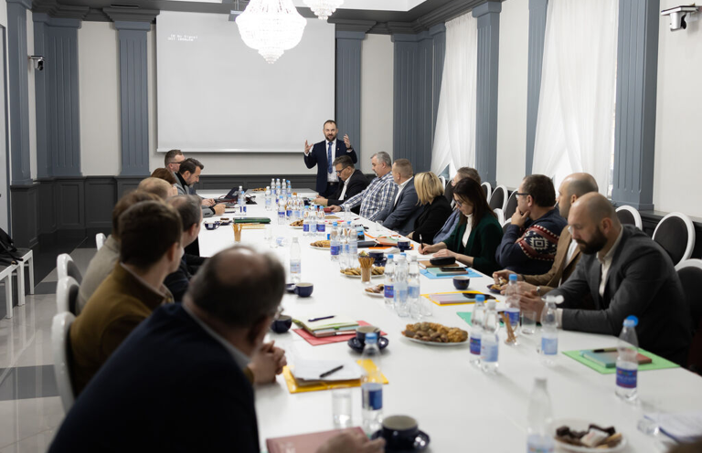 Sala konferencyjna spotkanie z przedsiębiorcami, wiele osób siedzących przy stole, słucha wykładu mężczyzny w garniturze