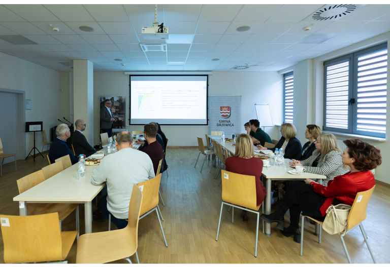Sala konferencyjna w Gminie Mniszków, kilka osób siedzi przy stołach słucha wykładu mężczyzny w garniturze, w tle ekran z obrazem z projektora