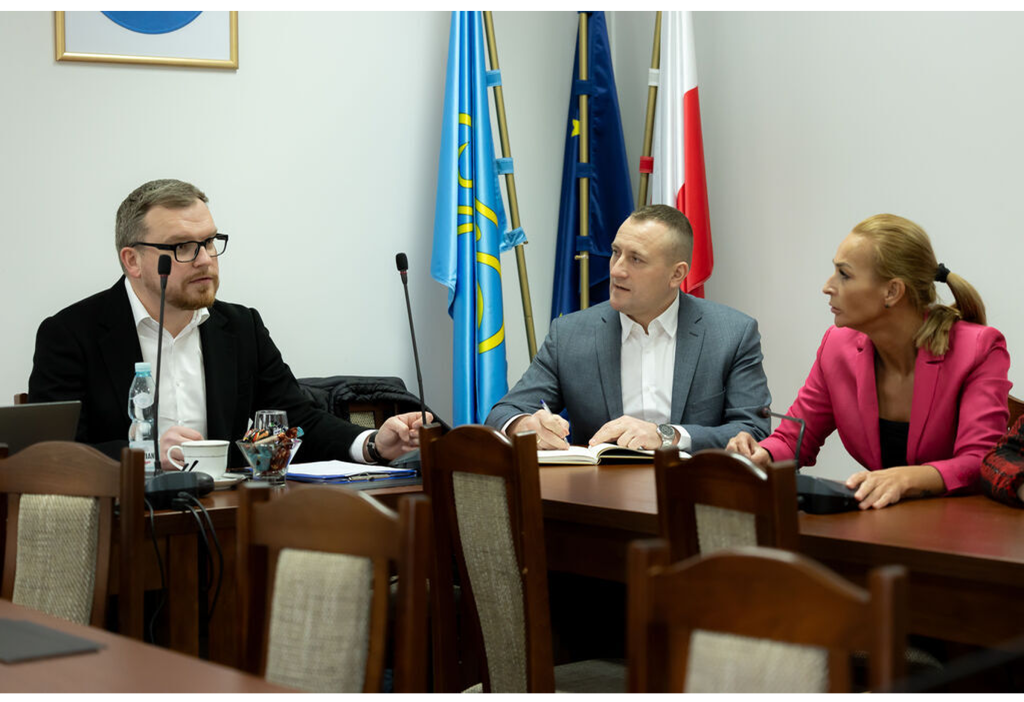 Kilka osób siedzi przy stole, na sali konferencyjnej urzędu gminy, w tle sztandary.