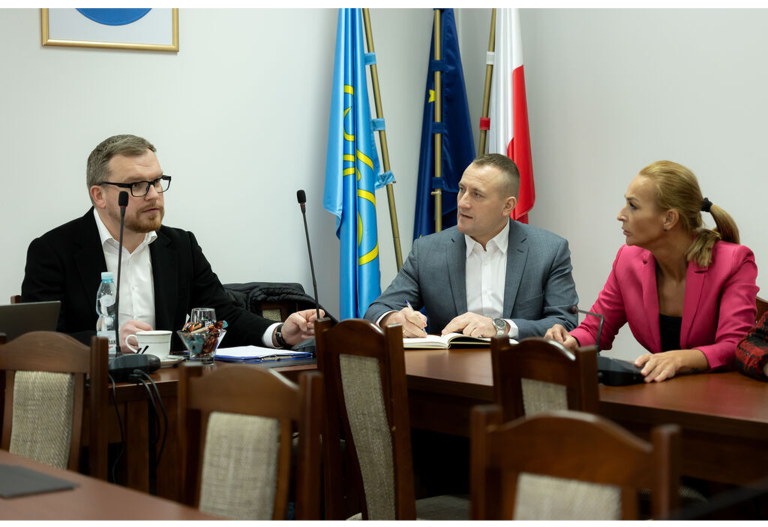 Kilka osób siedzi przy stole, na sali konferencyjnej urzędu gminy, w tle sztandary.